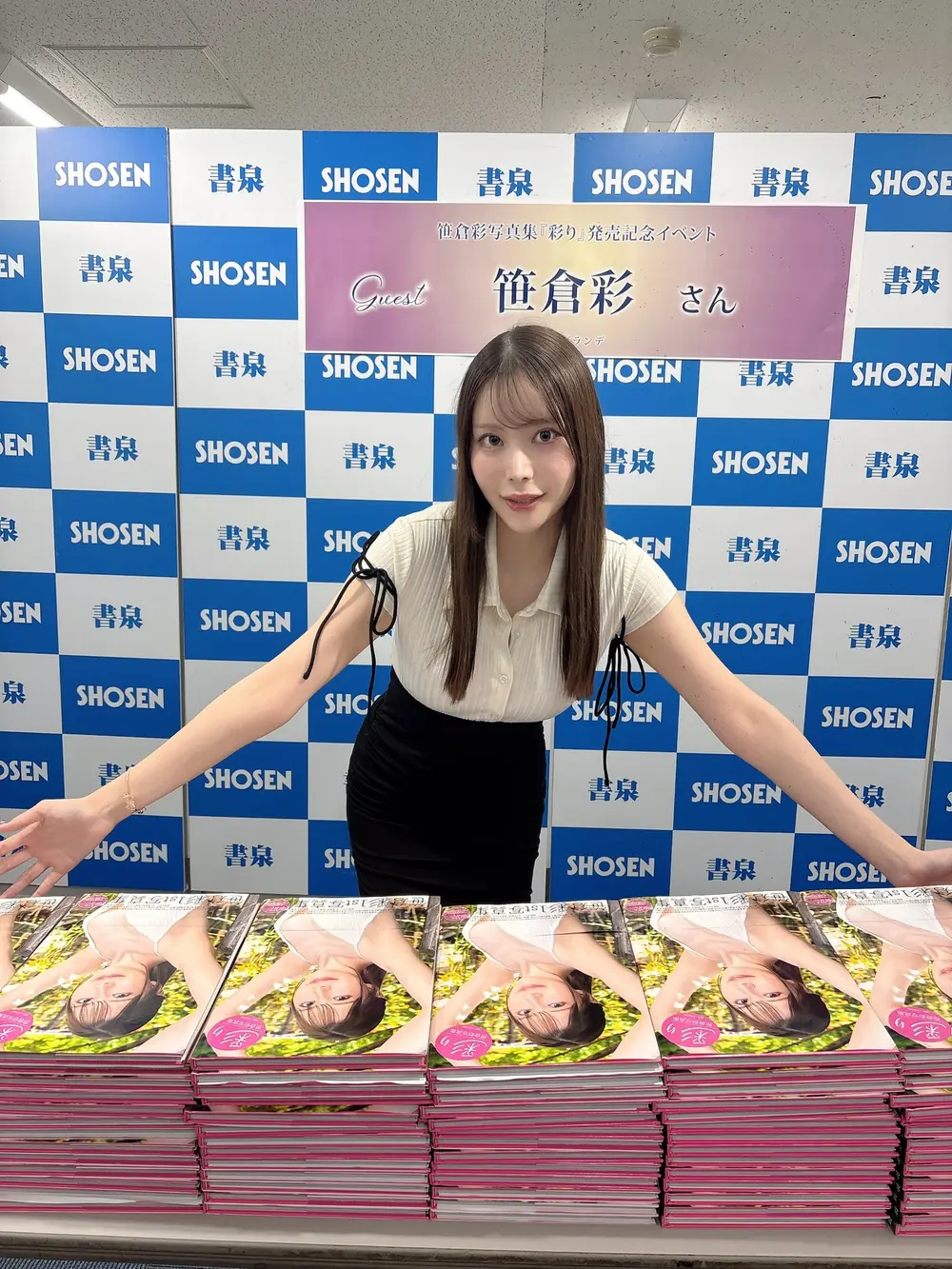 Aya Sasakura สวมเสื้อสีขาวและกระโปรงสีดำยืนอยู่หน้าโต๊ะหนังสือภาพในงานเปิดตัว