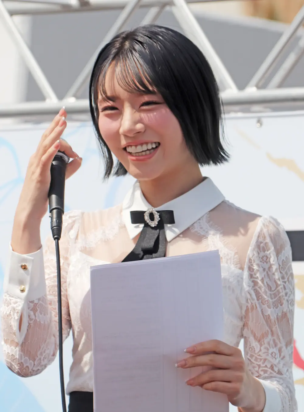 Saika Kawakita ยิ้มขณะถือไมโครโฟนและกระดาษบนเวที