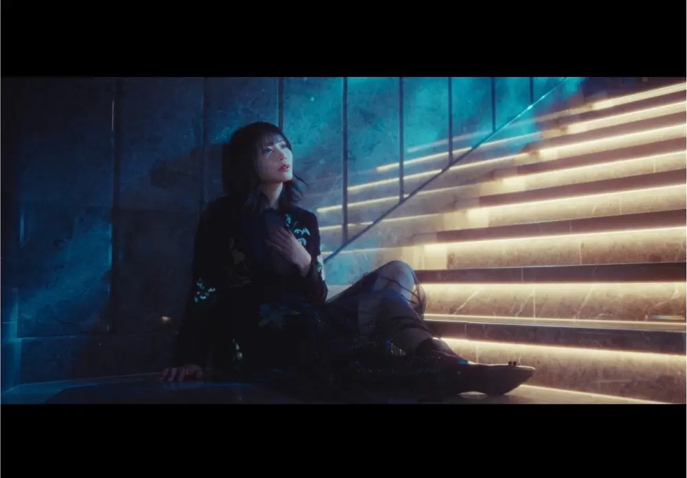 Saika Kawakita นั่งอยู่ริมบันไดที่มีแสงสีในภาพจากมิวสิกวิดีโอ