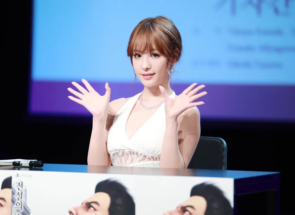 Kana Momonogi สวมชุดสีขาวและยกมือทักทายขณะนั่งบนเวที