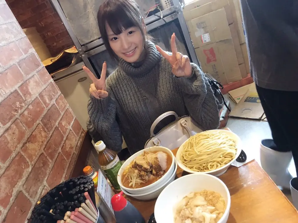 Kana Momonogi สวมเสื้อไหมพรมสีเทาและชูสองนิ้วทั้งสองข้างหน้าอาหารบนโต๊ะ