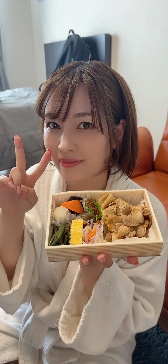 Saki Okuda ยิ้มและถือกล่องอาหารพร้อมชูสองนิ้วในชุดสีขาว