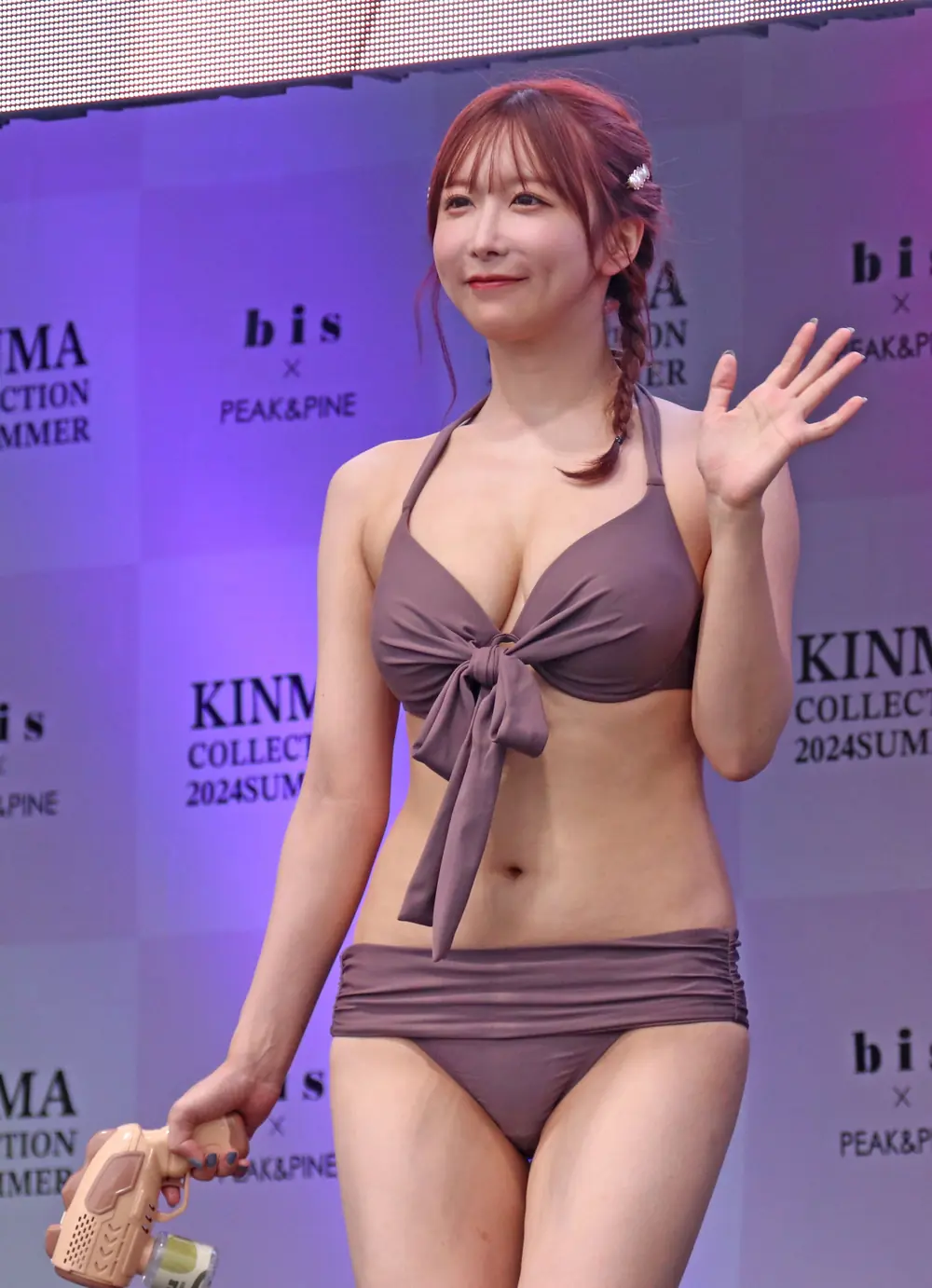 Unpai สวมชุดว่ายน้ำสีน้ำตาลอมม่วง ยกมือทักทายและถือปืนฉีดน้ำบนเวที
