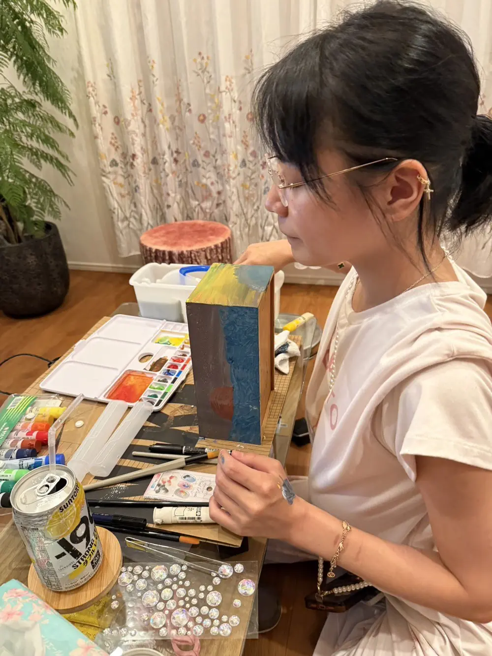 Yu Tano สวมเสื้อสีอ่อนนั่งวาดภาพที่โต๊ะพร้อมอุปกรณ์ระบายสี