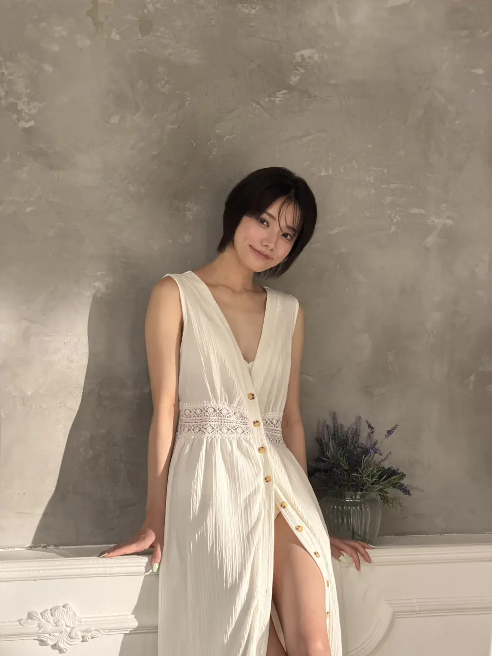 Rena Miyashita สวมเดรสยาวสีขาวและยืนพิงผนังสีเทา