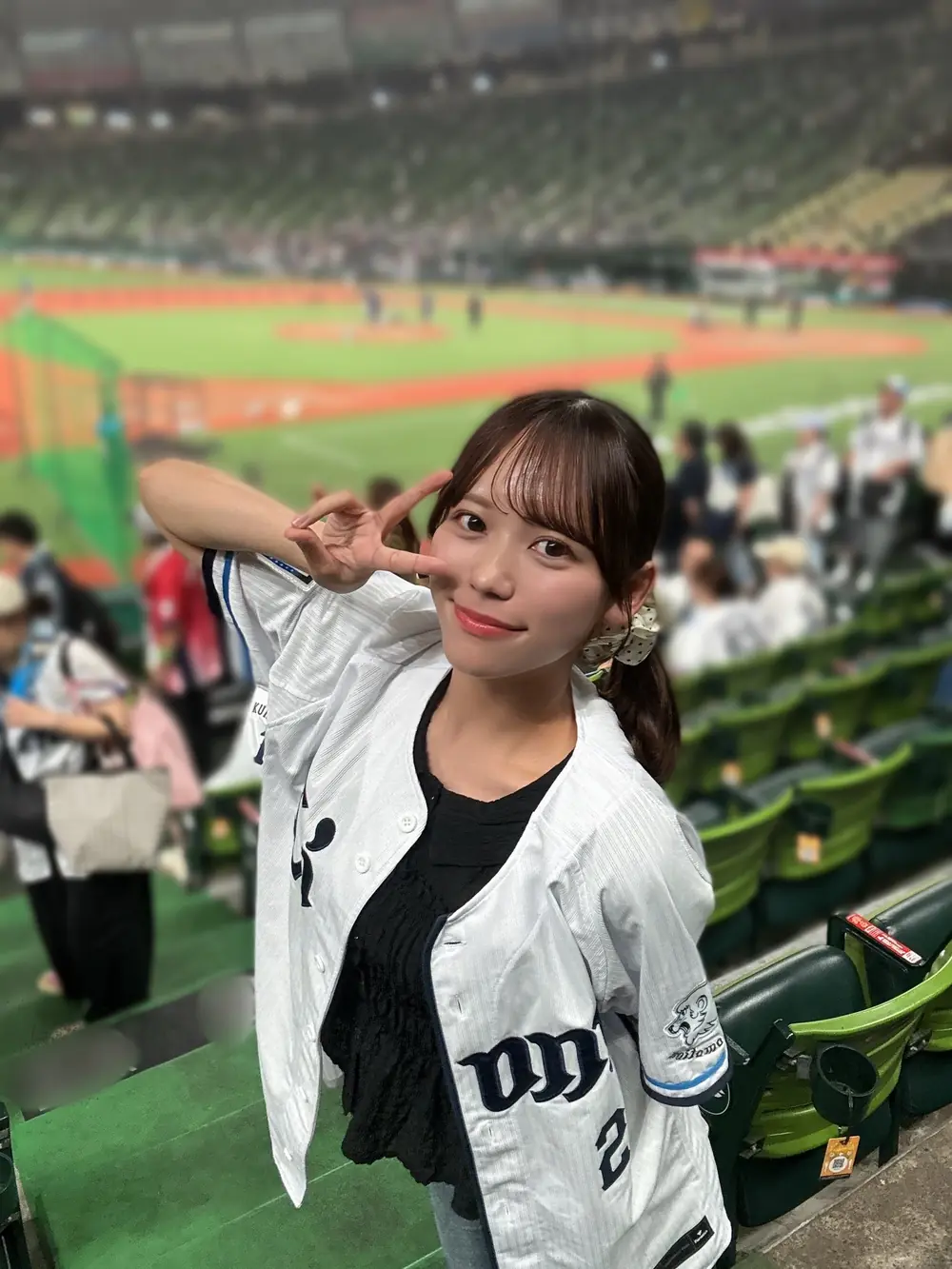 Mai Onodera สวมเสื้อทีมเบสบอลสีขาวยืนบนอัฒจันทร์ในสนาม