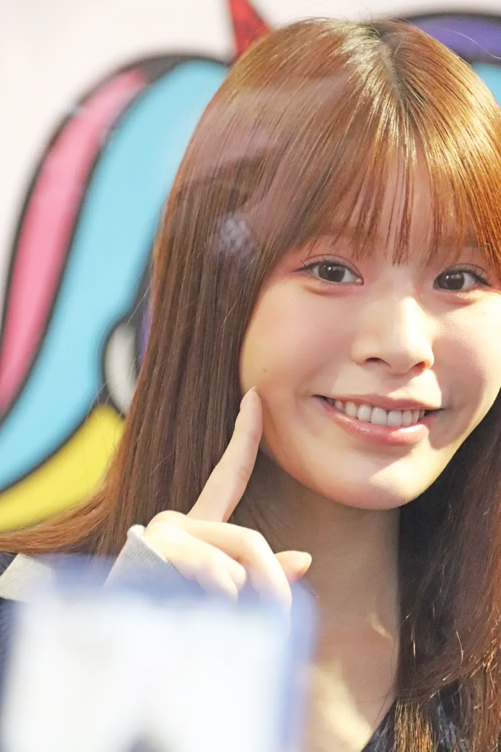 Nano Ogasawara ยิ้มและใช้นิ้วชี้แตะแก้มหน้าฉากสีสันสดใส
