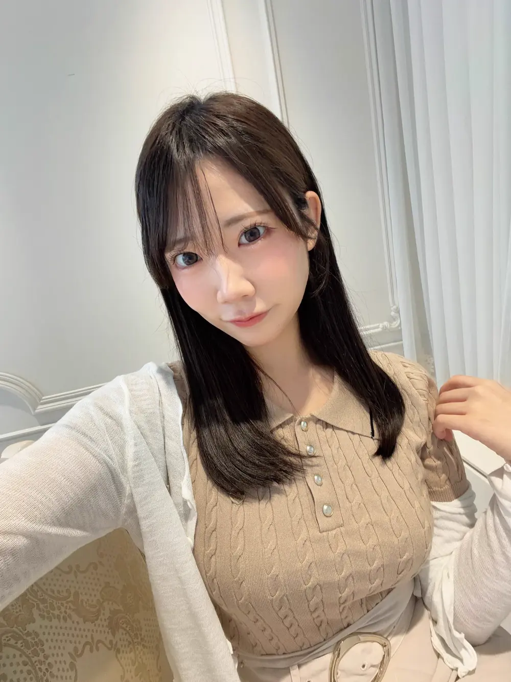 Yuno Sakura สวมเสื้อถักสีเบจและเสื้อคลุมสีขาวถ่ายภาพในห้อง