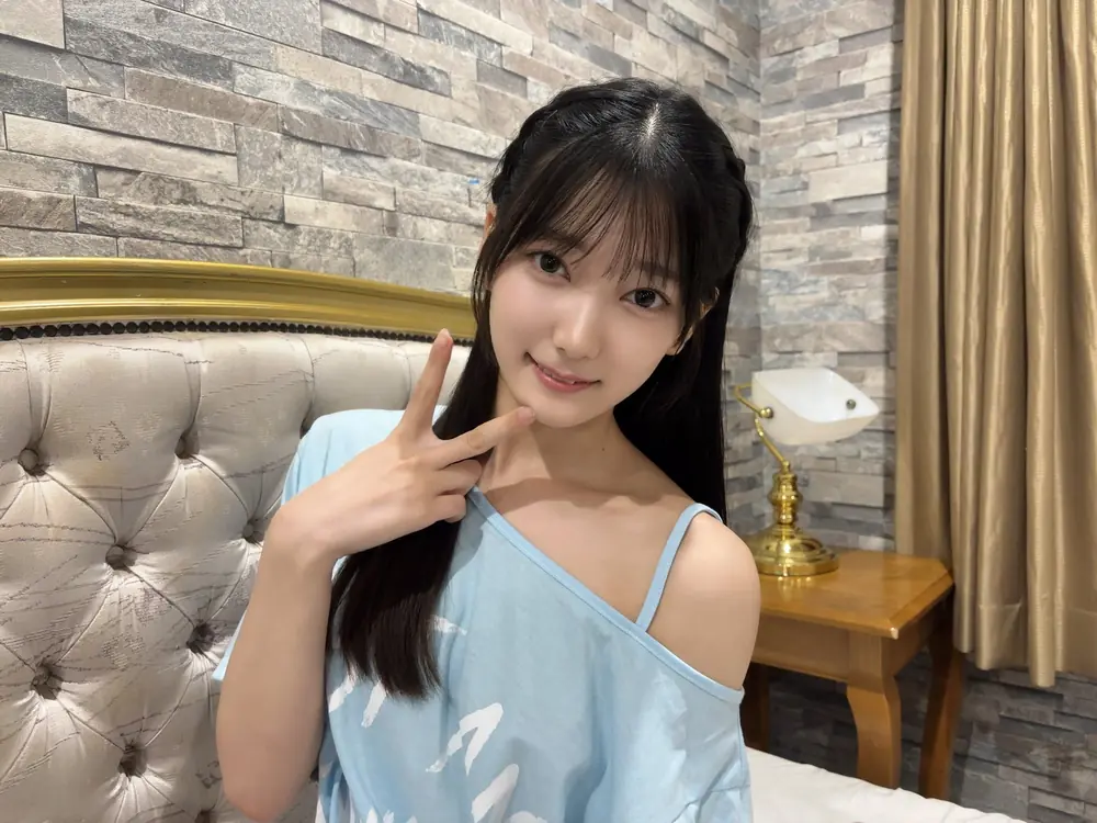 Nanoka Hatsumi สวมเสื้อสีฟ้าและชูสองนิ้วข้างใบหน้า