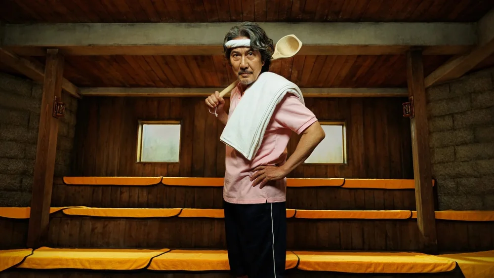 ชายสูงวัยในเสื้อสีชมพูยืนอยู่ในห้องซาวน่า มีผ้าขนหนูพาดไหล่และถือกระบวย