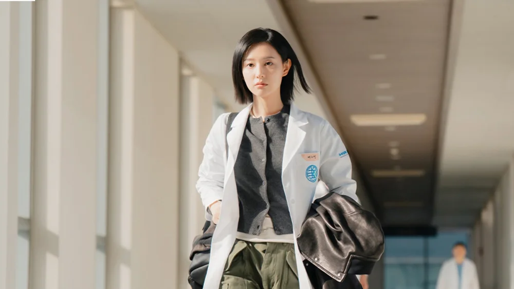 ภาพหญิงสาวใส่ชุดกาวน์เดินอยู่ในทางเดินโรงพยาบาล สะพายเสื้อหนังสีดำ
