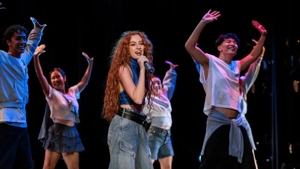 นักแสดงนำร้องเพลงบนเวทีโดยมีนักเต้นประกอบฉากอยู่ด้านหลัง