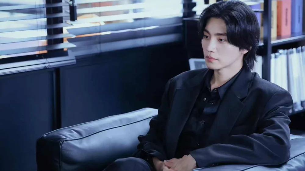 นักแสดงชายในชุดสูทนั่งบนโซฟาในออฟฟิศที่มีม่านบังแดด