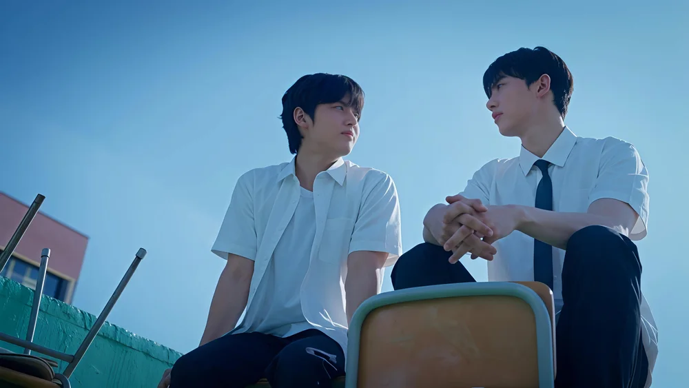 ชายสองคนนั่งบนดาดฟ้าใต้ท้องฟ้าสีฟ้า มองหน้ากัน
